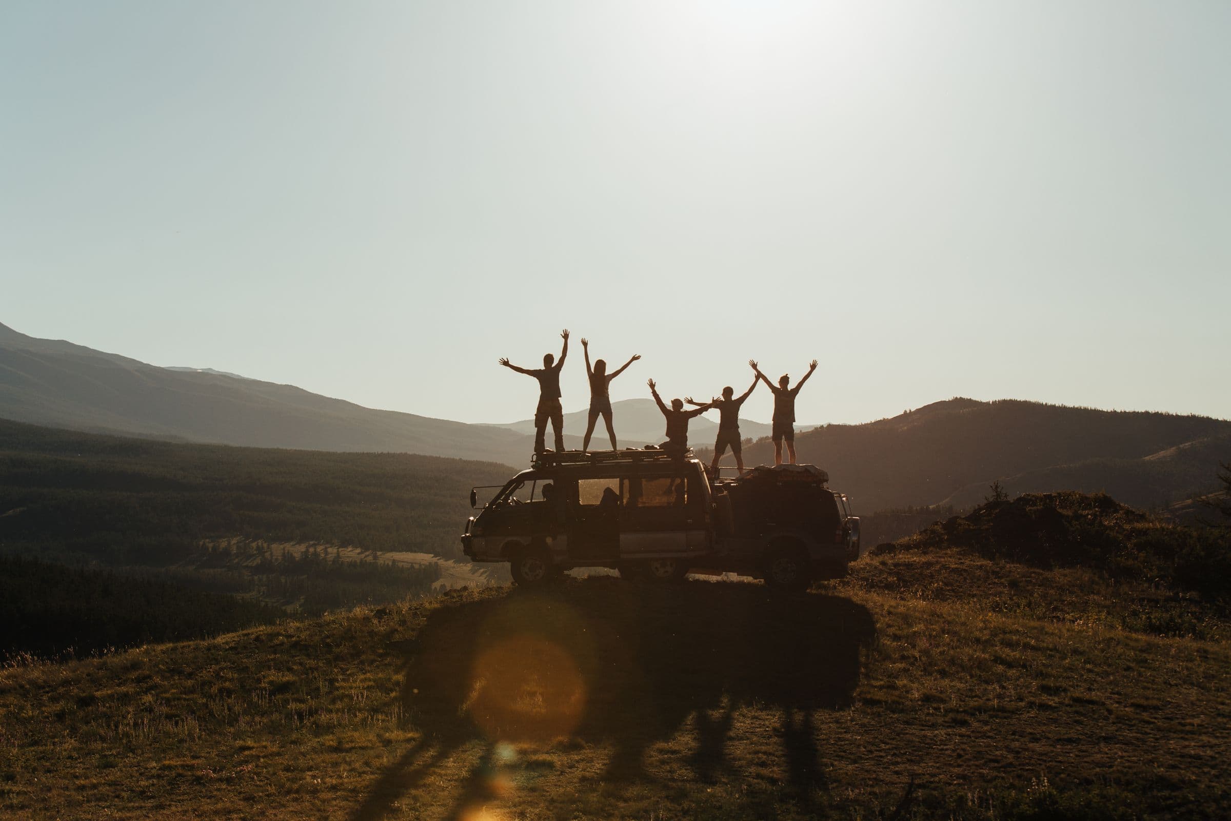 Five people raise their arms on top of a van in a mountain landscape.